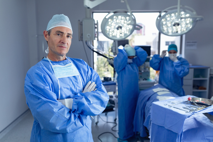 Portrait of Caucasian male surgeon standing with arm crossed while diverse surgeons performing surgery in operation room while surgeons getting ready for operation at hospital